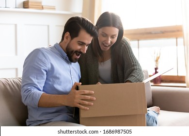 Surprised Young Clients Looking Inside Cardboard Box, Satisfied With Ordered Item. Happy Smiling Family Couple Opening Carton Parcel In Living Room, Amazed By Fast Delivery High Quality Service.