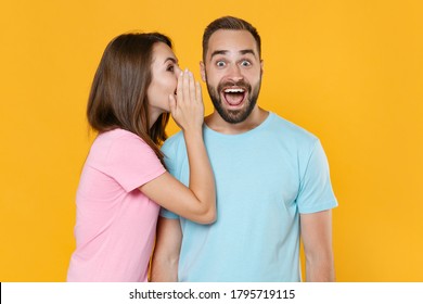 Surprised Young Couple Friends Guy Girl In Blue Pink T-shirts Isolated On Yellow Background Studio Portrait. People Lifestyle Concept. Mock Up Copy Space. Whispering Secret Behind Hand, Sharing News.