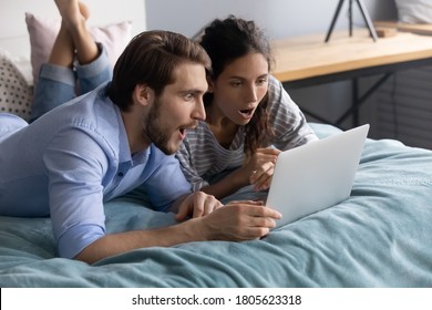 Surprised young family couple looking at computer screen, lying on bed. Shocked millennial man and woman reading email with unbelievable news, feeling astonished together in bedroom, omg reaction.