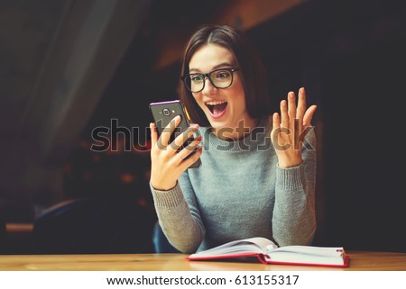 Surprised young female blogger chatting with best friends in social network on mobile phone while received shocking good news using wifi zone and reading interesting book indoors after work