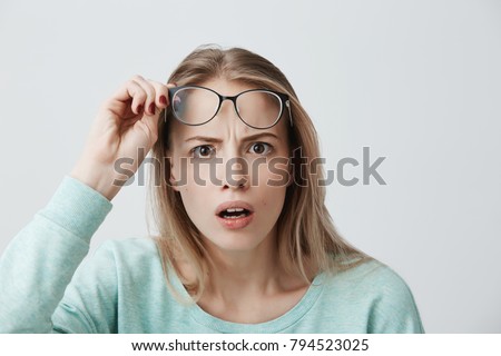 Surprised young female model with long blonde hair, wears glasses and blue long-sleeved shirt, looks with terror at camera as notices something unexpected, hears bad news, isolated against gray wall