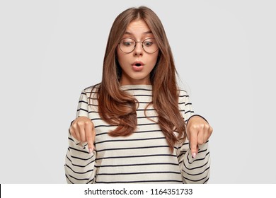 Surprised young lovely female teenager opens mouth widely, has startled look, points down with stupefied expression, sees something unbelievable, isolated over white background. Omg, look there!