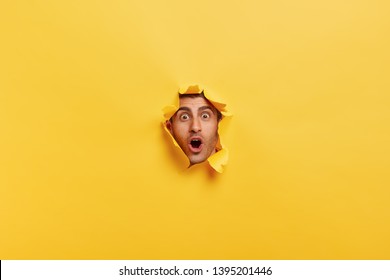 Surprised Young Man With Bristle Keeps Mouth Widely Opened, Stares Through Torn Paper Wall, Shows Only Face, Expresses Wonder And Disbelief, Poses Against Yellow Background. Facial Expressions