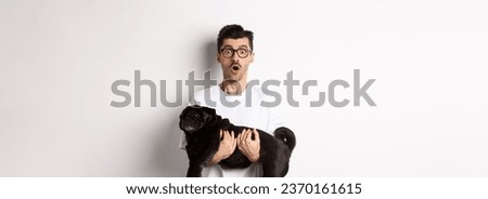 Surprised young man in glasses holding cute black pug, dog owner staring at camera with impressed face, saying wow, standing over white background.