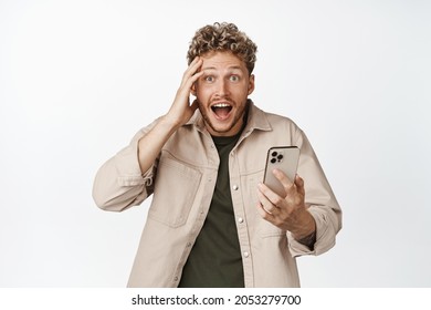 Surprised young man holding mobile phone, looking in disbelief at camera, amazed by smth on smartphone app, standing against white background
