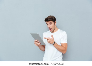 Surprised young man holding and pointing on tablet over grey background