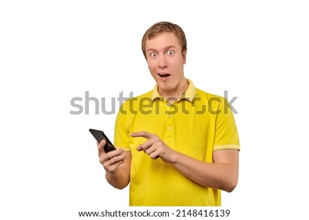Surprised young man with smartphone isolated on white background. Funny astonished man in yellow T-shirt getting push notifications using smart phone. Stunned happy guy receiving good news