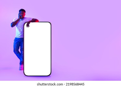 Surprised Young  Man Standing Near Giant Cellphone With Empty White Screen In Neon Light, Mockup For Mobile App, Website, Advertisement Design. Smartphone Template With Free Space