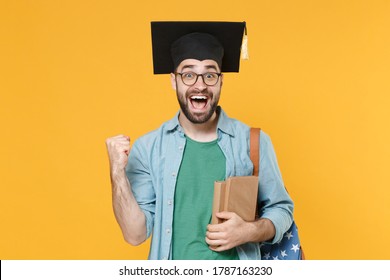 Surprised young man student in graduation cap glasses backpack hold books isolated on yellow background. Education in high school university college concept. Mock up copy space. Doing winner gesture