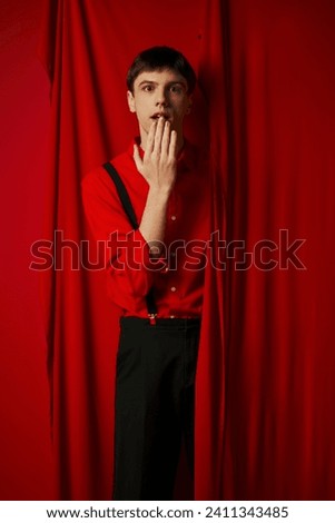surprised young man in suspenders smiling and covering mouth near red vibrant curtain, merriment