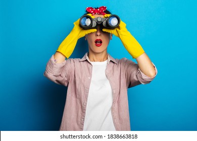 Surprised young woman in cleaning gloves looking through binoculars on blue background.