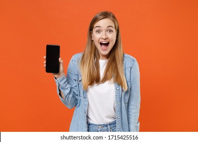 Surprised young woman girl in casual denim clothes posing isolated on orange wall background studio portrait. People lifestyle concept. Mock up copy space. Hold mobile phone with blank empty screen