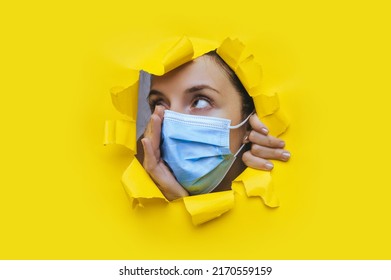 Surprised Young Woman Looks Through A Torn Hole In Yellow Paper And Adjusts A Protective Medical Mask. The Concept Of Protection Measures Against Pandemic Fatigue, Quarantine And Coronavirus Covid-19.