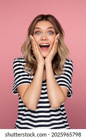 Surprised young woman with open mouth holding face in palm hands, isolated over pink background. Excited girl dressed in striped t-shirt wow emotion looking in camera. Surprise and shock concept.