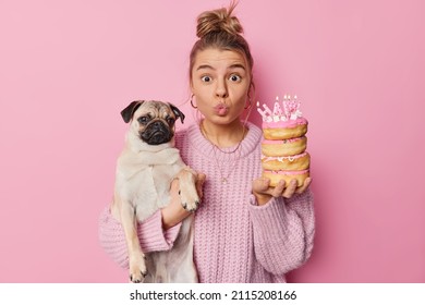 Surprised young woman pet owner keeps lips folded looks with wonder at camera holds birthday doughnuts with candles and pug dog celebrates bday of favorite pet poses against rosy background.