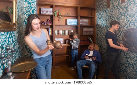 Surprised Young Woman Spending Time With Her Husband And Teen Sons In Quest Room Stylized As Old Library, Examining Ancient Kettle To Find Clue