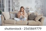 Surprised young woman is using mobile phone while sitting on comfortable sofa at home, reading shocking news in message or email, female looking at screen with amazement