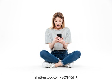 Surprised young woman using smartphone while sitting cross-legged on floor isolated