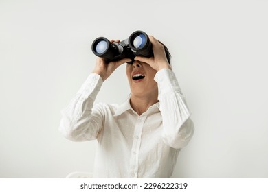 Surprised young woman in a white shirt looks through binoculars on a white background. - Powered by Shutterstock