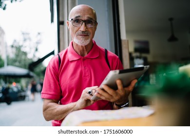 Surprized Aged Male In Bright Pink Shirt And Eyeglasses With Raised Brows Browsing Internet On Tablet While Sitting At Table In Street Cafe And Looking At Camera