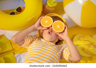 Surprized Child Holding Slices Of Orange Fruit Like Sunglasses. Kid Wearing Striped Yellow T-shirt Lying On Beach Towel. Healthy Eating And Summer Vacation Concept