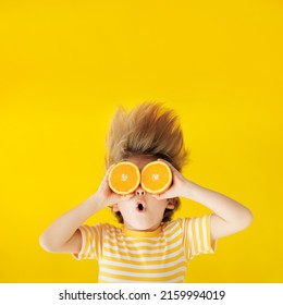 Surprized Child Holding Slices Of Orange Fruit Like Sunglasses. Kid Wearing Striped Yellow T-shirt Against Blue Paper Background. Healthy Eating And Summer Vacation Concept