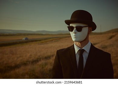 A surreal gentleman in a black suit, a bowler hat and a white mask stands looking away against the backdrop of a valley. Copy space.