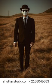 Surreal masked gentleman. Full-length portrait of a man in a black suit, bowler hat and white mask, standing in a field.