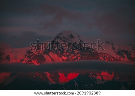 Surreal mountain landscape with great snowy mountains lit by dawn sun among low clouds. Fantasy alpine scenery with high mountain pinnacle at sunset or at sunrise. Big glacier on top in neon red light