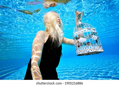 Surreal Portrait Of A Blonde Girl Underwater In An Outdoor Pool. She Poses In A Black Dress And Holds A White Check. Her Face Is Covered By Her Hair. Faceless. Bright Colors. Fashion Photography.