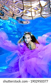 A Surreal Portrait Of A Teenage Girl. She Looks Into The Camera And Poses Underwater At The Bottom Of An Outdoor Pool In A Purple Dress And A Wreath Of Flowers. Underwater Fashion Photography.