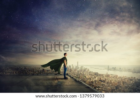 Surreal view of confident superhero with cape stands on the rooftop looking over city horizon. Ambition and business success concept. Leadership hero power, motivation and inner strength symbol.