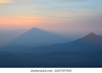 Surreal view of a perfect triangular shadow cast by Volcán de Acatenango during sunrise in Guatemala. The misty atmosphere and pastel sky create a magical and serene moment high above the landscape
