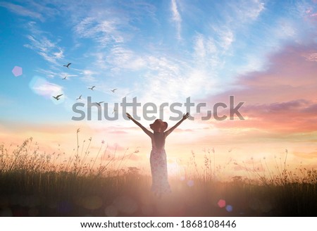 Surrender and praising concept: Silhouette of healthy Christian woman raised hands at meadow sunset background