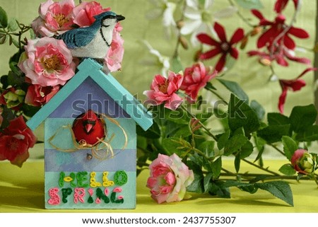 surrounded by artificial flowers on a yellow-green background there is a decorative birdhouse with birds and with a colorful inscription on it hello spring
