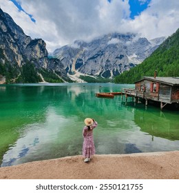 Surrounded by towering mountains, a girl in a floral dress stands by an emerald lake, enjoying the tranquil view. A rustic cabin and a small boat Lago Di Braies Dolomites Italy - Powered by Shutterstock