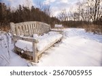 Surrounded by trees in the forest, an antique, wooden bench sits blanketed in snow on a sunny, winter day in Houghton, Michigan.