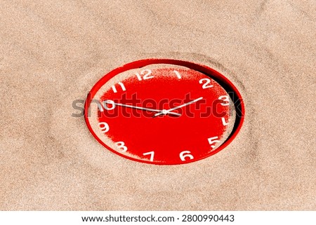 surrounded by an unbroken surface of beach sand, a red clock showing ten past ten appears slowly swallowed by the shoreline landscape under strong sunlight