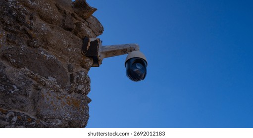 Surveillance camera fixed on an old stone wall under bright blue sky, concept of security and control.