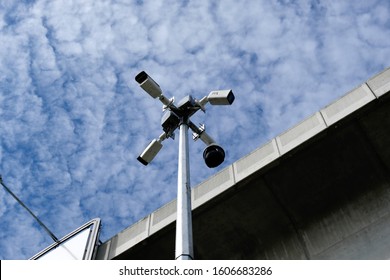 Surveillance Camera At A Street Intersection Monitoring Road Traffic In All Directions. CCTV Mounted On A Pole To Monitor Traffic Conditions On The Streets Below. Cloudy Skies With Flyover Backdrop.