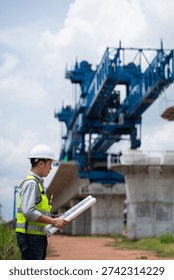 The survey team is operating a theodolite while referencing road construction plans. Civil engineers are taking measurements with surveying equipment at the construction site.