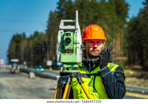 Surveyor engineer with equipment\
(theodolite or total positioning station) on the construction site\
of the road or building with construction machinery\
background
