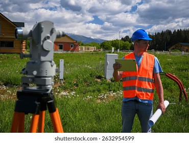 Surveyor using tablet and theodolite is working on construction site with houses and mountains in background.