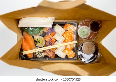 Sushi To Go Concept. Top View Of Takeaway Box With Sushi Rolls And Various Sauce Cups In Brown Paper Bag.