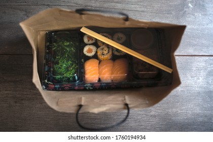 Sushi To Go Concept. Top View Of Takeaway Box With Sushi Rolls And Chopsticks In Brown Paper Bag On Wood Floor. Maki. Sashimi. Salmon. Tuna. Wakame. Asian.Japanese. Paper Bag.Take-away Japanese Food.