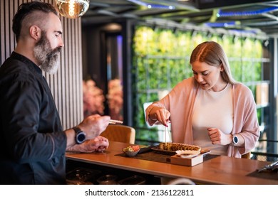 Sushi master is teaching young woman how to use chopsticks at sushi restaurant.