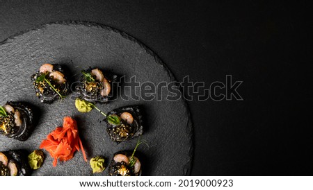 Sushi roll with nori, black rice, crab meat, cucumber, avocado, smoked salmon mousse, oar caviar, masago, shrimp cocktail and edible gold leaf with red ginger and wasabi on black table and background.