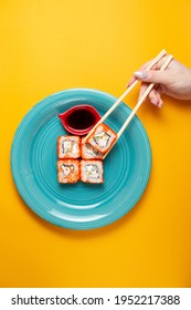 Sushi rolls with salmon on a black with soy sauce and a woman's hand with chopsticks on a yellow background