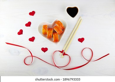 Sushi for Valentines Day in a heart-shaped plate on white wooden background. Sticks with a red ribbon, decorations-hearts. Top view