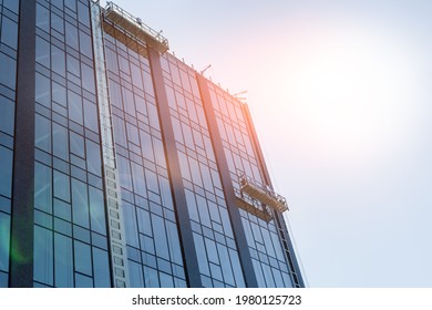 Suspended construction craddle near wall of hightower residentaial building with glass metal modern facade construction site. Engineering urban city development background. Sun shining flare on sky